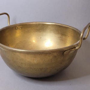 Könnte beinhalten: Eine Vintage-Messingschüssel mit zwei gebogenen Griffen. Die Schüssel hat eine abgerundete Form und eine glatte, polierte Oberfläche. Die Griffe sind an den Seiten der Schüssel befestigt. Dieser Küchenartikel wird wahrscheinlich zum Kochen oder Servieren verwendet.