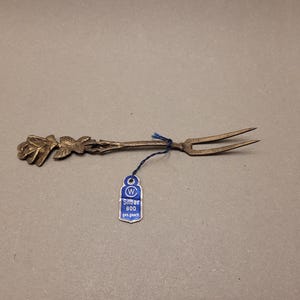 May include: A silver-toned serving fork with a floral design on the handle. The fork has two tines and is attached to a blue tag with the text "Silber 800". The fork is likely used for serving appetizers or desserts.