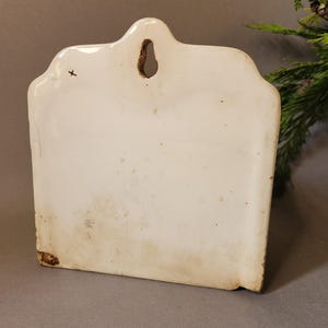 Puede incluir: Un bolsillo de pared de cer&aacute;mica blanco roto vintage con un borde superior decorativo y un agujero para colgar. El bolsillo rectangular tiene un aspecto ligeramente envejecido con algo de decoloraci&oacute;n y una peque&ntilde;a cruz.