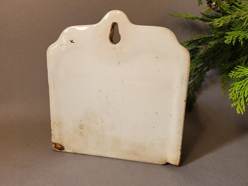 Puede incluir: Un bolsillo de pared de cer&aacute;mica blanco roto vintage con un borde superior decorativo y un agujero para colgar. El bolsillo rectangular tiene un aspecto ligeramente envejecido con algo de decoloraci&oacute;n y una peque&ntilde;a cruz.