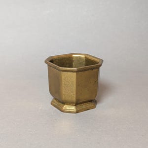 Könnte beinhalten: Ein kleines, achteckiges Gefäß aus Messing mit einer goldfarbenen Metalloberfläche. Der Behälter verfügt über eine weite, offene Oberseite mit einem definierten Rand, glatte vertikale Seiten und einen gestuften Boden.
