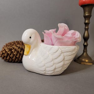 Peut inclure: Un pot en céramique blanc en forme de canard avec un bec jaune et des yeux noirs, rempli de fleurs roses. Un cône de pin brun et un bougeoir en laiton avec une bougie rouge sont en arrière-plan.