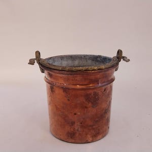 May include: A small copper bucket with a brass rim and two brass handles. The bucket has a worn, aged appearance.