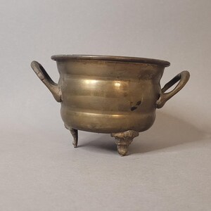 Peut inclure: Un petit bol en laiton antique avec deux poignées et trois pieds décoratifs. Le bol a une forme arrondie et un bord légèrement évasé. Le laiton présente une patine chaude et vieillie. Peut être utilisé à des fins décoratives.