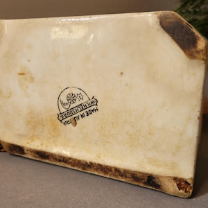 Puede incluir: Objeto de cer&aacute;mica color crema con forma rectangular, que muestra signos de edad y desgaste. La base y los bordes presentan decoloraci&oacute;n marr&oacute;n. La parte inferior tiene un sello negro que dice "MADE IN AUSTRIA WILHELMSBURG".
