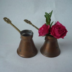 Può includere: Due piccole caffettiere di rame con manici in ottone. Le caffettiere sono riempite con rose rosa e foglie verdi.
