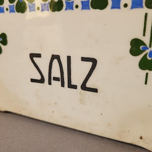 Puede incluir: Un recipiente de cer&aacute;mica blanco con la palabra "SALZ" en letras negras. El recipiente est&aacute; decorado con patrones florales verdes y azules. El dise&ntilde;o incluye motivos en forma de tr&eacute;bol y un borde de cuadrados azules y formas verdes alternas.