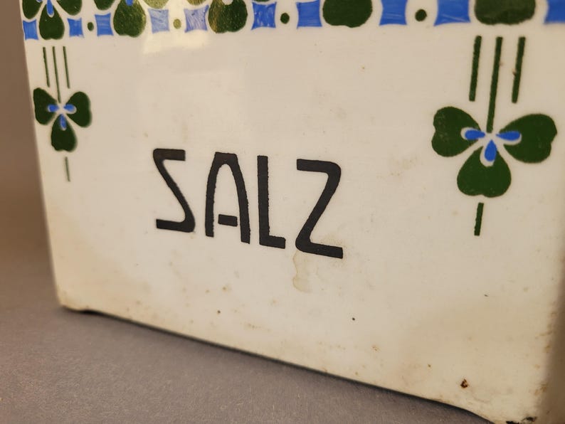 Puede incluir: Un recipiente de cer&aacute;mica blanco con la palabra "SALZ" en letras negras. El recipiente est&aacute; decorado con patrones florales verdes y azules. El dise&ntilde;o incluye motivos en forma de tr&eacute;bol y un borde de cuadrados azules y formas verdes alternas.