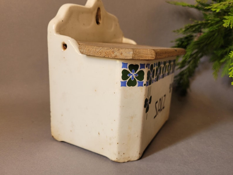 Puede incluir: Salero antiguo de cer&aacute;mica blanca con tapa de madera. La caja presenta un dise&ntilde;o floral en azul, verde y negro. La palabra "SALT" est&aacute; escrita en negro en el lateral. Un art&iacute;culo de decoraci&oacute;n de cocina vintage.