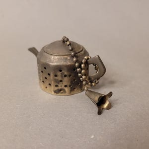 Könnte beinhalten: Vintage Tee-Ei in Form einer kleinen Teekanne in Silberoptik. Das Tee-Ei hat einen Deckel, einen Ausguss und einen Griff. Eine Kette aus kleinen Perlen verbindet das Tee-Ei mit einem glockenförmigen Ende. Die Teekanne hat kleine Löcher zum Ziehen von Tee.