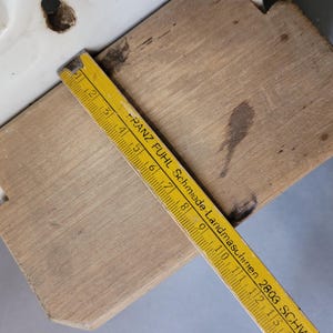 Puede incluir: Una tabla de cortar de madera con un asa de cer&aacute;mica blanca se muestra con una regla amarilla que mide su longitud. La regla tiene marcas negras y el texto "FRANZ FUHL Schmiede-Landmaschinen 2803 SCHW".
