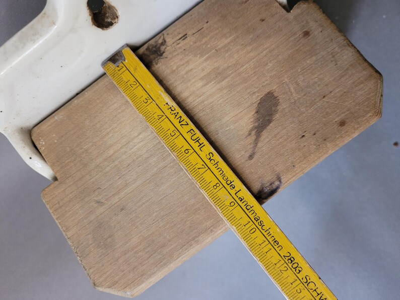 Puede incluir: Una tabla de cortar de madera con un asa de cer&aacute;mica blanca se muestra con una regla amarilla que mide su longitud. La regla tiene marcas negras y el texto "FRANZ FUHL Schmiede-Landmaschinen 2803 SCHW".