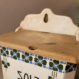 Puede incluir: Una salera blanca vintage con tapa de madera. La caja presenta un borde decorativo de dise&ntilde;os florales verdes y azules. La palabra "SALT" es visible en la parte delantera. La caja tiene una parte superior decorativa con un agujero para colgar.