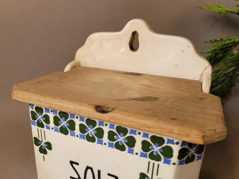 Puede incluir: Una salera blanca vintage con tapa de madera. La caja presenta un borde decorativo de dise&ntilde;os florales verdes y azules. La palabra "SALT" es visible en la parte delantera. La caja tiene una parte superior decorativa con un agujero para colgar.