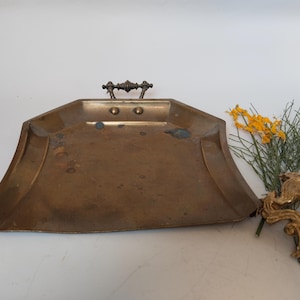 Peut inclure: Une pelle à poussière en laiton avec une base plate et large et une petite poignée en métal décorative fixée sur le bord arrière. La surface métallique présente des signes d'usure avec des taches de patine sombre et un fini terne.