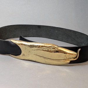May include: A black leather belt featuring a large, polished gold-tone metal buckle shaped like a stylized snake head. The buckle has a textured, hammered finish and a curved, elongated form that secures the belt strap with a smooth, dark surface.