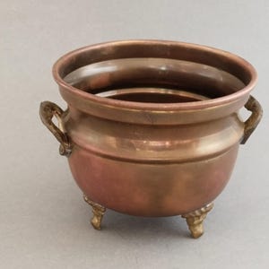Puede incluir: Una olla redonda de cobre con un acabado metálico liso, que cuenta con dos pequeñas asas laterales y tres patas cortas decorativas. El recipiente tiene una abertura ancha y un cuerpo redondeado, mostrando un tono marrón rojizo cálido.