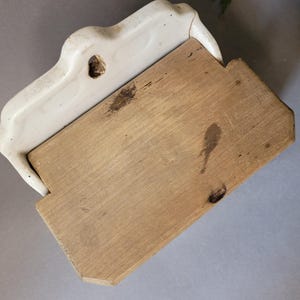 Puede incluir: Un jabonera vintage de cer&aacute;mica blanquecina con base de madera. La jabonera tiene un dise&ntilde;o curvo y un agujero para el drenaje. La base de madera es rectangular con vetas naturales y algunas manchas oscuras.