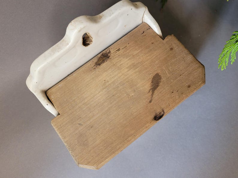 Puede incluir: Un jabonera vintage de cer&aacute;mica blanquecina con base de madera. La jabonera tiene un dise&ntilde;o curvo y un agujero para el drenaje. La base de madera es rectangular con vetas naturales y algunas manchas oscuras.