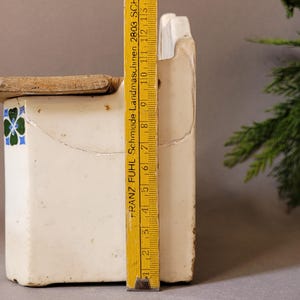 Puede incluir: Un recipiente de cer&aacute;mica blanca vintage con tapa de madera, con un dise&ntilde;o floral decorativo en azul y verde. Una regla amarilla, marcada con medidas, se coloca junto al recipiente. El recipiente mide aproximadamente 15 cm de alto.