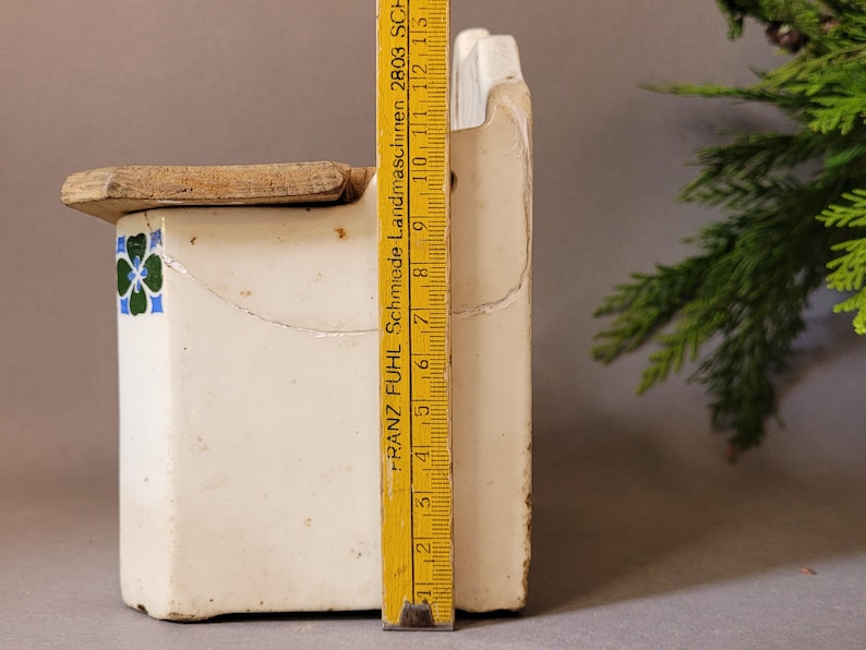 Puede incluir: Un recipiente de cer&aacute;mica blanca vintage con tapa de madera, con un dise&ntilde;o floral decorativo en azul y verde. Una regla amarilla, marcada con medidas, se coloca junto al recipiente. El recipiente mide aproximadamente 15 cm de alto.