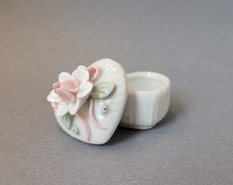 May include: A white heart-shaped porcelain trinket box with a lid featuring a sculpted white and pale pink flower, green leaves, and a pink ribbon detail. The base of the container is plain white with a ribbed texture along the exterior sides.