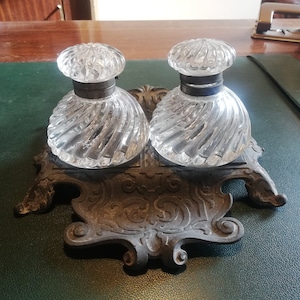 Op de afbeelding: Twee heldere glazen inktpotten met sierlijke zilveren deksels, die op een decoratieve metalen standaard staan met ingewikkelde ontwerpen. De standaard heeft een donkerbruine afwerking.