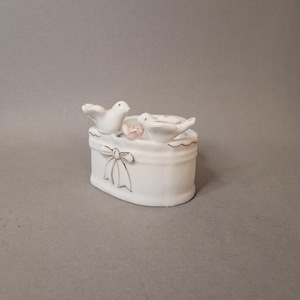 May include: A white ceramic trinket box with two doves perched on top. The box features a bow design with gold accents and a small pink flower. The overall design is delicate and decorative.