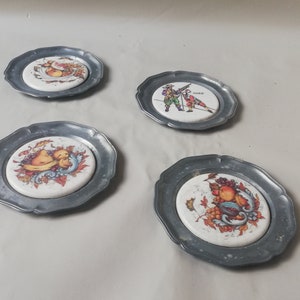 May include: Four vintage pewter coasters with decorative ceramic inserts. Each coaster features a different colorful fruit or figure design. The coasters have a scalloped edge and are set against a neutral background.