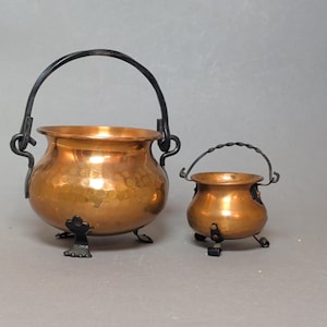 Pode incluir: Dois potes de cobre martelado com alças de metal escuro e três pés pequenos. O pote maior possui uma alça arqueada lisa, enquanto o menor tem uma alça de metal torcido. Ambos os recipientes têm um corpo arredondado e abertura larga.