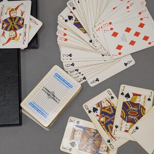 Può includere: Un mazzo di carte da gioco con una scatola nera. È visibile una carta jolly, insieme a un ventaglio di carte che mostrano vari semi e numeri. Un mazzo mostra un retro blu con motivi e il testo E. Piatnik & Sohne Wien.