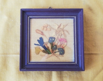 May include: A framed pressed flower artwork featuring a selection of dried flowers and leaves in shades of brown, pink, blue, and green. The flowers are arranged in a bouquet shape and are displayed against a beige background.