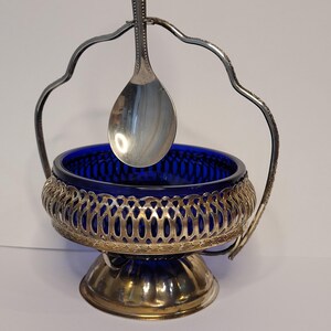May include: An antique silver-plated sugar bowl with a cobalt blue glass insert. The bowl has a decorative pierced metalwork design around the rim and sits on a pedestal base. A silver spoon hangs from the handle.
