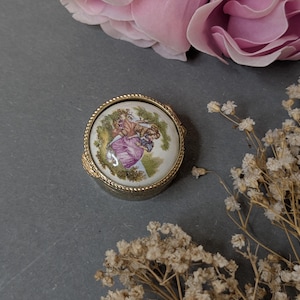 May include: A round metal trinket box with a gold-toned, textured rim and a circular ceramic lid insert. The lid features a colourful painted scene of two figures in period clothing standing in a landscape with green foliage and a white background.