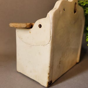 Puede incluir: Un especiero de cer&aacute;mica blanco roto vintage para colgar en la pared, con una peque&ntilde;a balda de madera. El especiero rectangular tiene un borde superior decorativo y muestra signos de antig&uuml;edad, como grietas y decoloraci&oacute;n. Una peque&ntilde;a balda de madera sobresale por delante.