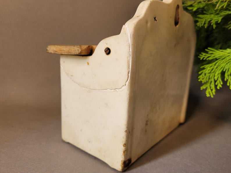 Puede incluir: Un especiero de cer&aacute;mica blanco roto vintage para colgar en la pared, con una peque&ntilde;a balda de madera. El especiero rectangular tiene un borde superior decorativo y muestra signos de antig&uuml;edad, como grietas y decoloraci&oacute;n. Una peque&ntilde;a balda de madera sobresale por delante.