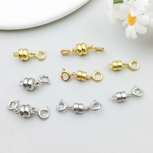 Könnte beinhalten: Eine Sammlung gold- und silberfarbener Magnetverschlüsse für Schmuck. Die Verschlüsse haben ein zylindrisches Design mit Magnetverschluss und kleinen Schlaufen zum Befestigen an Schmuckketten oder -schnüren. Die Verschlüsse werden vor weißem Hintergrund gezeigt.