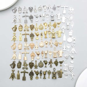 Puede incluir: Una colección de dijes de metal en tonos plata, oro y bronce, con diversos diseños temáticos de vino y bebidas como uvas, sacacorchos, botellas, copas y barriles, dispuestos en una cuadrícula sobre una superficie de color gris claro.