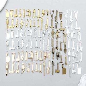 May include: A collection of metal charms shaped like various hand tools, including hammers, saws, wrenches, screwdrivers, axes, and rulers. The charms are finished in gold, silver, and bronze tones and are arranged in a grid pattern on a light surface.
