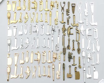 May include: A collection of metal charms shaped like various hand tools, including hammers, saws, wrenches, screwdrivers, axes, and rulers. The charms are finished in gold, silver, and bronze tones and are arranged in a grid pattern on a light surface.
