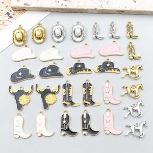 Puede incluir: Una colección de dijes de metal con diseños de temática western que incluyen sombreros de vaquero, botas, caballos y cráneos de toro. Los dijes tienen acabados en esmalte dorado, plateado, negro, blanco y rosa, con una pequeña argolla.
