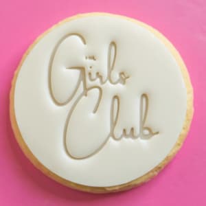 Peut inclure: Un biscuit rond avec une garniture blanche portant l'inscription "Girl Club" en cursive. Le biscuit est posé sur un fond rose vif. Le bord du biscuit est de couleur marron clair.