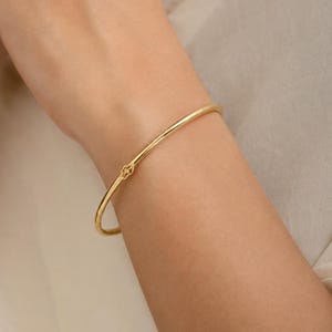 May include: A thin, polished gold bangle bracelet featuring a small, intricate, four-petaled floral or clover-shaped clasp detail centered on the smooth, circular band. The bracelet has a bright, reflective metallic finish and a simple design.