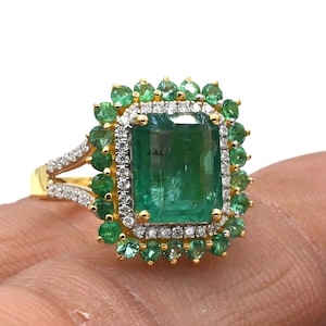 Puede incluir: Un anillo de oro con una gran esmeralda rodeada de esmeraldas y diamantes más pequeños.