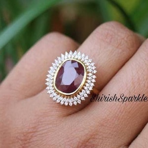 May include: A gold ring featuring a large, oval-cut, deep red gemstone surrounded by a halo of small, white gemstones. The ring is set against a blurred green background. The text "ShirishSparkle" is visible.