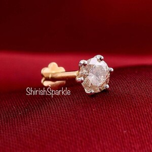 May include: A gold nose stud featuring a round, faceted, clear gemstone held in a six-prong setting. The post has a flat, flower-shaped back. The text ShirishSparkle is visible in white lettering on the left side of the jewelry piece.