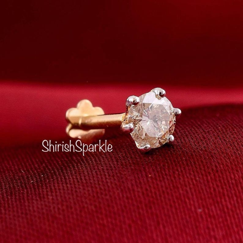 May include: A gold nose stud featuring a round, faceted, clear gemstone held in a six-prong setting. The post has a flat, flower-shaped back. The text ShirishSparkle is visible in white lettering on the left side of the jewelry piece.