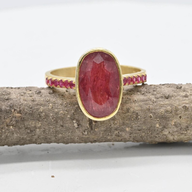 K&ouml;nnte beinhalten: Ein Goldring mit einem gro&szlig;en, oval geschliffenen, tiefroten Edelstein als Mittelpunkt. Die Ringschiene ist mit einer Reihe kleiner, runder, leuchtend roter Steine besetzt, die beidseitig eingelassen sind und ein symmetrisches Design bilden.