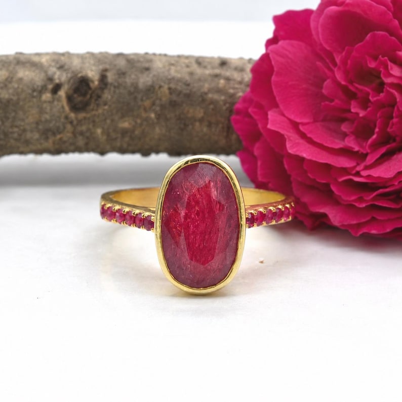 K&ouml;nnte beinhalten: Ein goldener Ring mit einem gro&szlig;en, ovalen, facettierten roten Edelstein in der Mitte, der von einer d&uuml;nnen Goldfassung umgeben ist. Die Ringschiene ist mit einer Reihe kleiner, runder, facettierter roter Steine an beiden Seiten besetzt.
