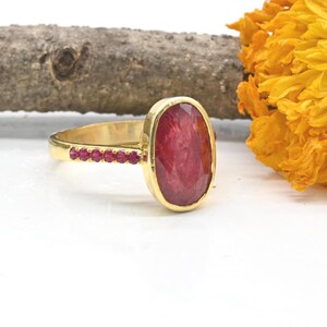 K&ouml;nnte beinhalten: Ein goldener Ring mit einem gro&szlig;en, ovalen, facettierten roten Edelstein als Herzst&uuml;ck. Die Ringschiene ist mit einer Reihe kleiner, runder, roter Steine besetzt, die seitlich eingelassen sind und ein stimmiges Gesamtbild ergeben.